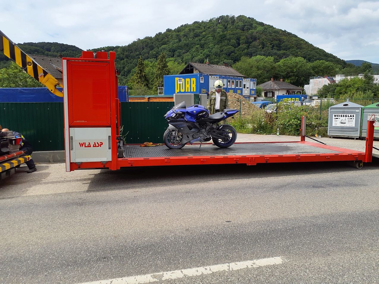 Ein blaues Motorrad wird auf einen roten Flachbettanhänger verladen. Ein Mann mit weißem Helm steht daneben. Hinter dem Anhänger befindet sich ein blaues Gebäude mit der Aufschrift "PIRR".