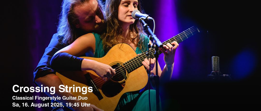 Eine Frau spielt Gitarre und singt in ein Mikrofon, während ein Mann hinter ihr steht und sanft ihre Schulter küsst. Die Bühne ist mit lila Lichtern beleuchtet.