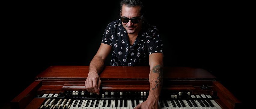 Ein Mann mit Sonnenbrille spielt Orgel. Er hat ein Tattoo am Arm und lächelt. Das Klavier ist aus Holz mit schwarzen und weißen Tasten.