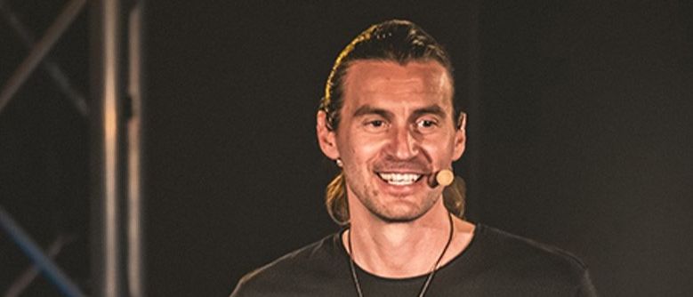 Ein Mann steht auf der Bühne, hält ein Buch mit dem Titel 'AUF DEM WEG ZU MIR' in den Händen, lächelt und gestikuliert. Er trägt ein schwarzes T-Shirt und hat ein Mikrofon am Hals.