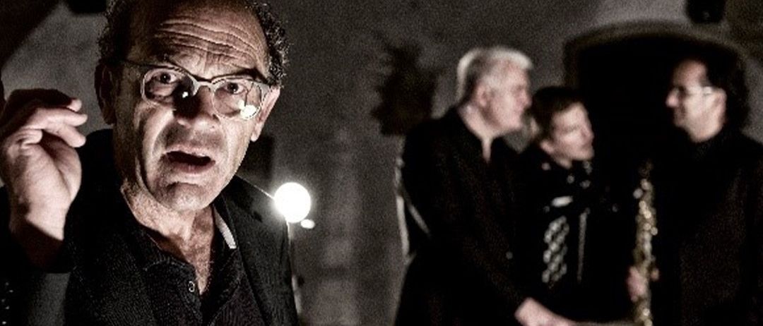 Ein älterer Mann mit Brille steht in einem schwach beleuchteten Raum und macht eine Handbewegung. Hinter ihm spielt ein Mann Saxophon. Zwei weitere schauen zu.