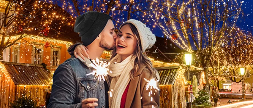 Ein Paar steht unter einem geschmückten Baum, lächelt und hält Funkenfeuerwerk in der Hand an einem verschneiten Abend. Hinter ihnen ist ein Gebäude mit Weihnachtslichtern beleuchtet.