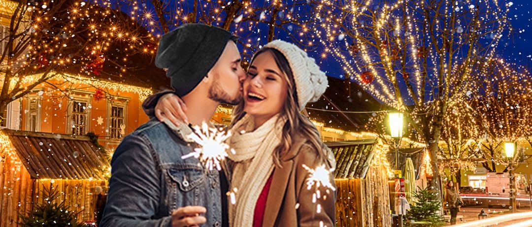 Ein Paar steht unter einem verschneiten Himmel, hält Wunderkerzen in den Händen und lächelt, während sie sich umarmen, mit Weihnachtslichtern, die den Hintergrund erhellen.