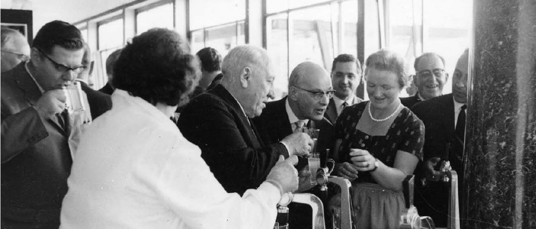 Ein altes Foto einer Gruppe von Menschen, einschließlich eines älteren Mannes in Anzug und Krawatte, einer Frau in Kleid und Halskette und anderen, die um eine Bar stehen, wahrscheinlich bei einer gesellschaftlichen Veranstaltung.