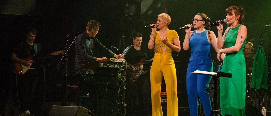 Eine Band tritt auf der Bühne auf, ein Mann spielt die Tastatur und drei Frauen singen in Mikrofone.