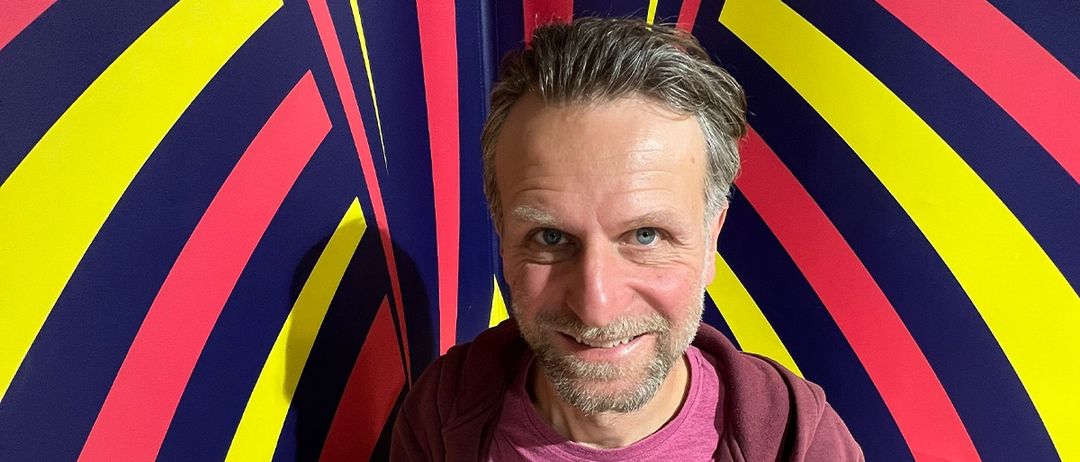 Ein Mann mit Bart und Schnurrbart steht vor einer farbenfrohen, abstrakten Wand. Er trägt eine bordeauxfarbene Kapuzenjacke und ein rosa T-Shirt und lächelt in die Kamera.