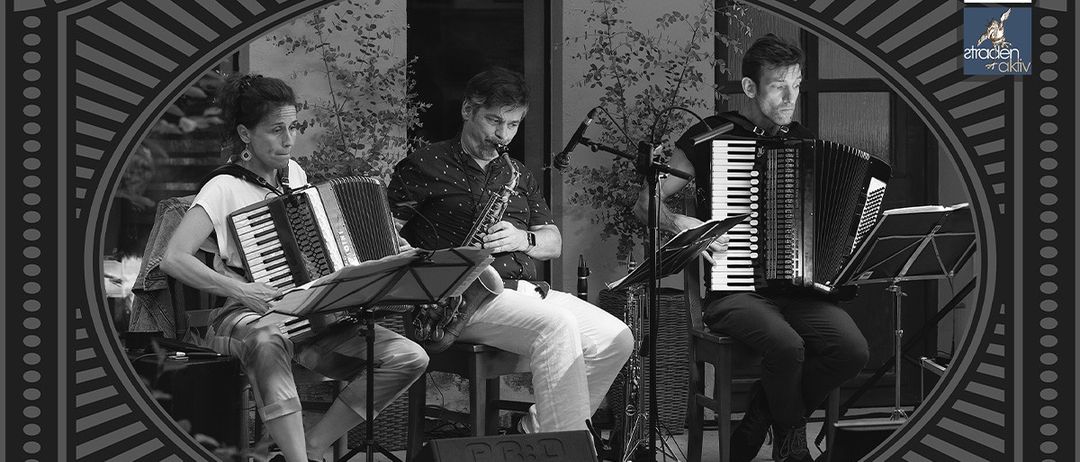 Drei Musiker spielen Instrumente bei einer Veranstaltung im Freien. Eine Frau spielt Akkordeon, ein Mann Saxophon und ein weiterer Mann Akkordeon. Sie sitzen und sind von Musikinstrumenten und Mikrofonen umgeben.