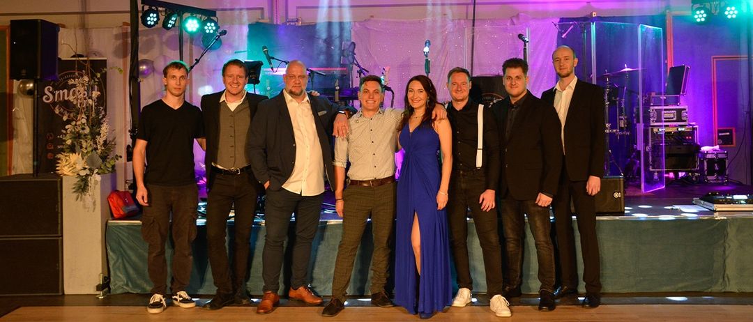 Eine Gruppe von formell gekleideten Menschen posiert für ein Foto auf einer Bühne mit lila Beleuchtung und Mikrofonen.