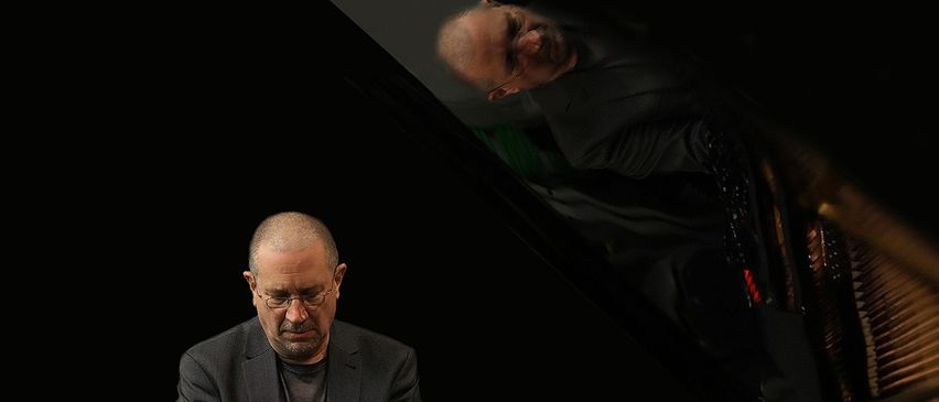 Ein Mann sitzt konzentriert an einem Klavier, seine Reflexion zeigt das Profil eines anderen Mannes über ihm. Er trägt eine Brille und einen grauen Anzug.