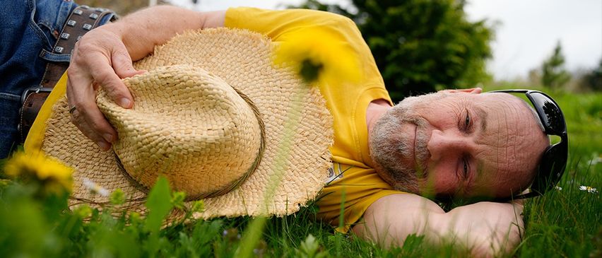 Ein älterer Mann in einem gelben T-Shirt liegt auf dem Gras, hält einen Strohhut und lächelt in die Kamera.