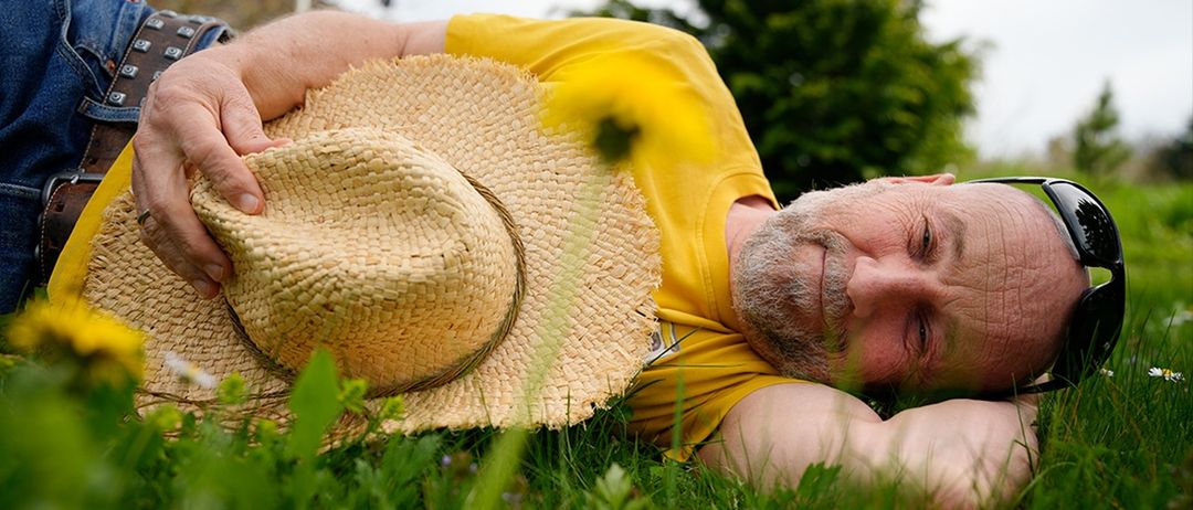 Ein älterer Mann in einem gelben T-Shirt liegt auf dem Gras, hält einen Strohhut und lächelt in die Kamera.