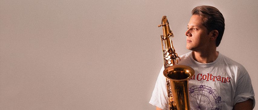 Ein Mann hält ein Saxophon dicht vor sein Gesicht und trägt ein weißes T-Shirt mit einem roten 'A'-Logo und einem Riesenrad-Design.