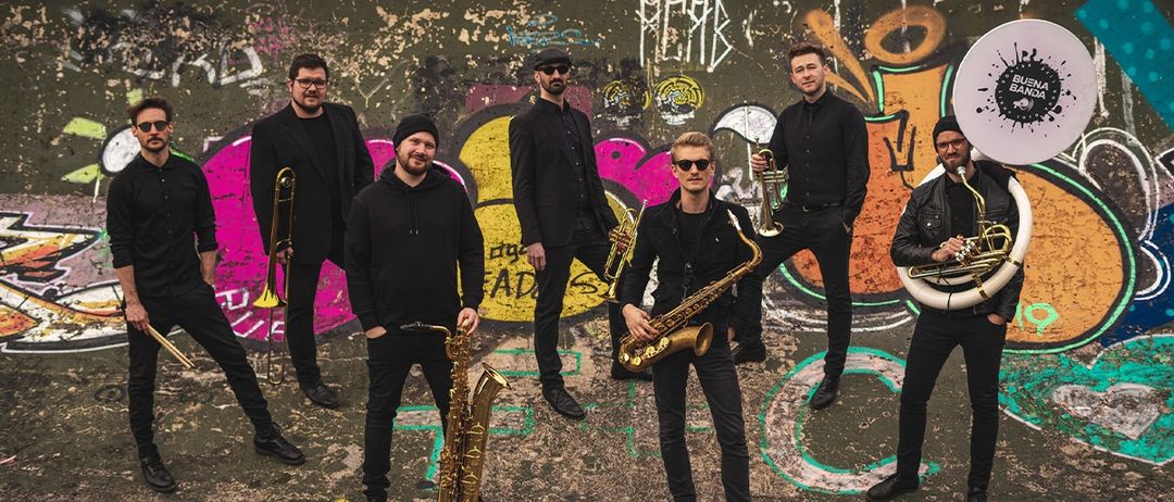 Eine Gruppe von Musikern steht vor einer mit Graffiti bedeckten Wand, jeder hält ein Blechblasinstrument und ist in Schwarz gekleidet.