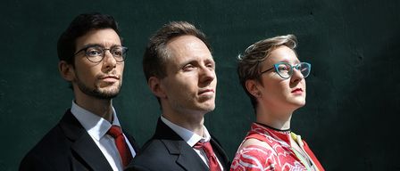 Drei Personen, alle in formeller Kleidung, stehen nebeneinander vor einer grünen Wand und schauen in verschiedene Richtungen. Der Mann auf der linken Seite hat einen Bart und eine Brille.