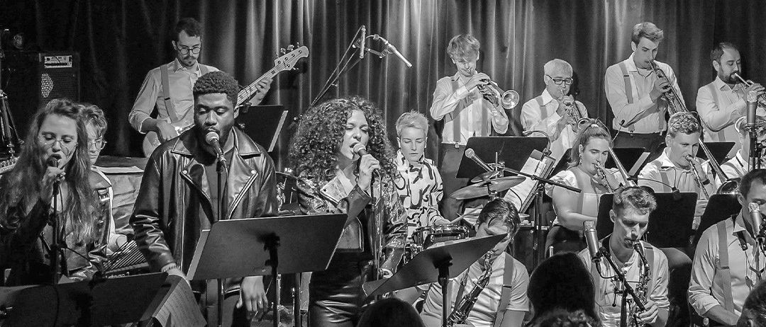 Ein Schwarz-Weiß-Foto einer Band, die auf der Bühne von Tube's auftritt, mit Musikern, die verschiedene Instrumente spielen, und einem Publikum, das zuschaut.