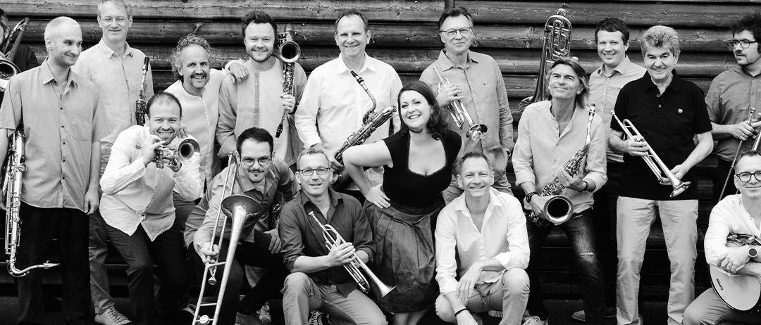 Ein Schwarz-Weiß-Foto zeigt eine große Gruppe von Musikern, die vor einer Holzwand posieren und jedes sein Instrument hält. Einige stehen, andere sitzen.