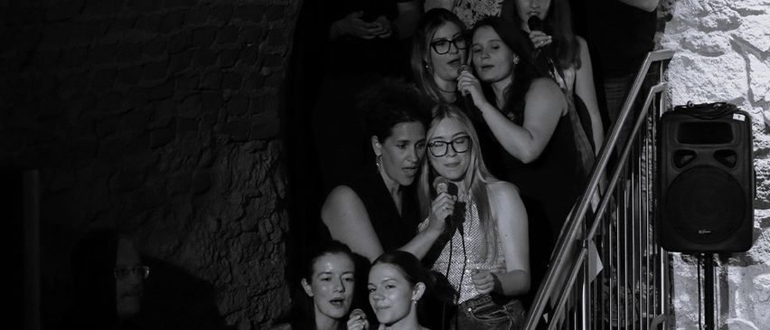 Eine Gruppe von Menschen steht in einem schwach beleuchteten Raum, einige halten Mikrofone. Einige stehen auf einer Treppe, und ein Lautsprecher ist an der Wand montiert.
