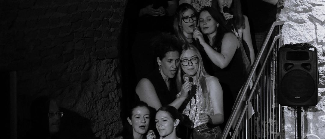 Eine Gruppe von Menschen steht in einem schwach beleuchteten Raum, einige halten Mikrofone. Einige stehen auf einer Treppe, und ein Lautsprecher ist an der Wand montiert.