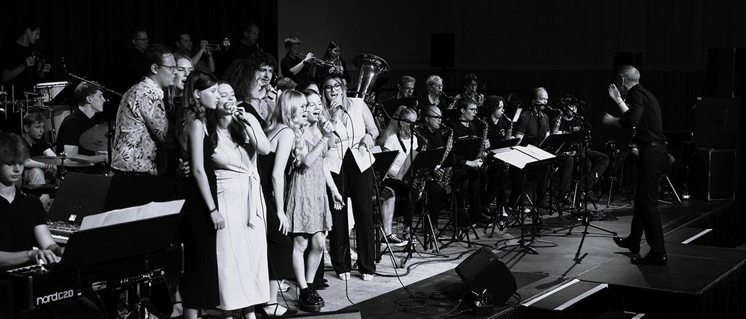 Eine Band tritt auf der Bühne auf, mit Sängern und Instrumentalisten, einschließlich Trompeten und Saxophonen. Die Bühne ist mit Mikrofonen, Lautsprechern und Notenständern ausgestattet.