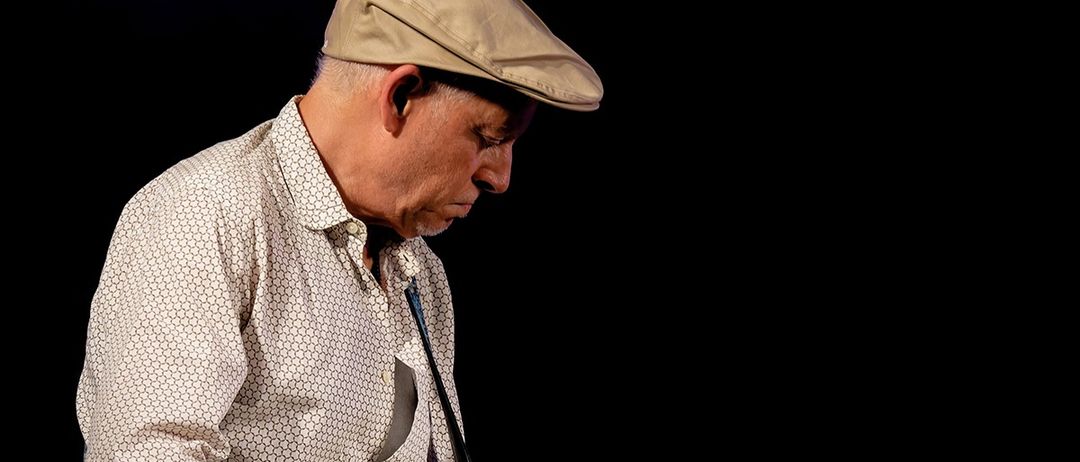 Ein älterer Mann mit beigem Cap und gepunktetem Hemd spielt Gitarre und schaut nach unten.