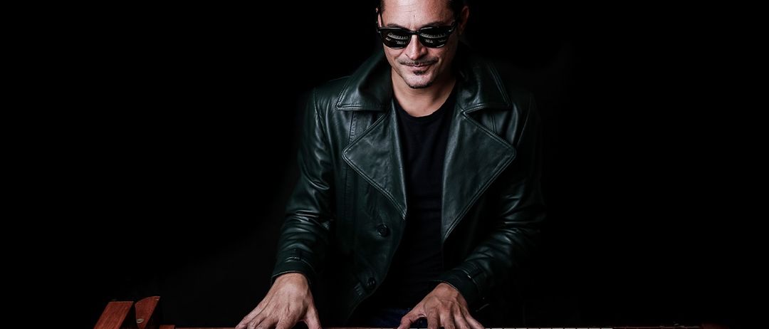 Ein Mann in einer schwarzen Lederjacke und Sonnenbrille spielt mit seinen Händen an einem elektrischen Klavier.