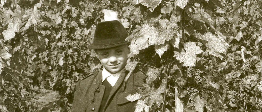 Ein Junge in Hut und Mantel steht zwischen Weinreben und lächelt in die Kamera. Das Bild ist in Schwarz-Weiß.