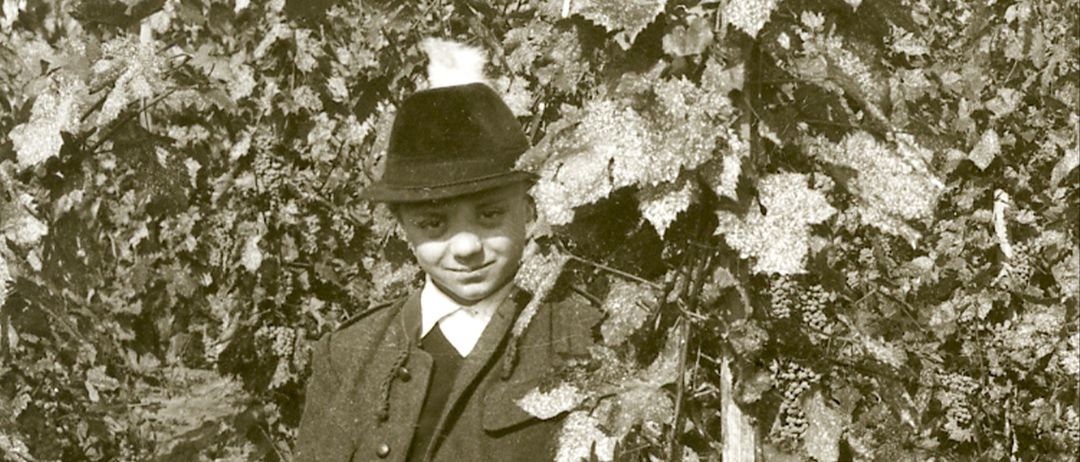 Ein Junge in Hut und Mantel steht zwischen Weinreben und lächelt in die Kamera. Das Bild ist in Schwarz-Weiß.