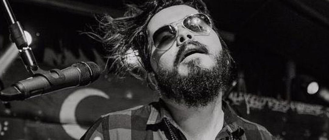 Ein Mann mit Bart und Sonnenbrille singt in ein Mikrofon. Der Hintergrund zeigt einen Halbmond mit Text.