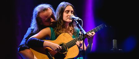A man and a woman are on stage. The woman is playing a guitar while singing into a microphone. The man has his head on her shoulder.