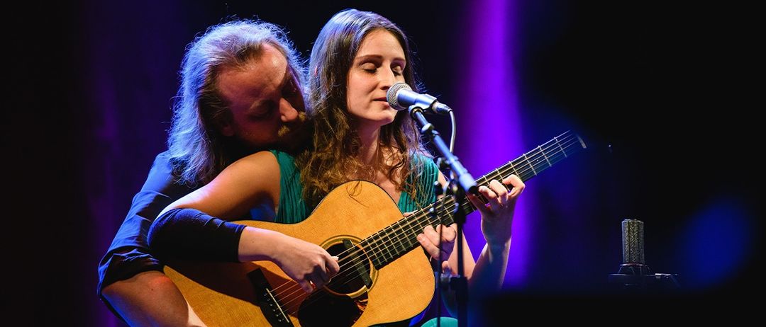 Ein Mann und eine Frau sind auf der Bühne. Die Frau spielt eine Gitarre und singt in ein Mikrofon. Der Mann hat seinen Kopf auf ihrer Schulter.
