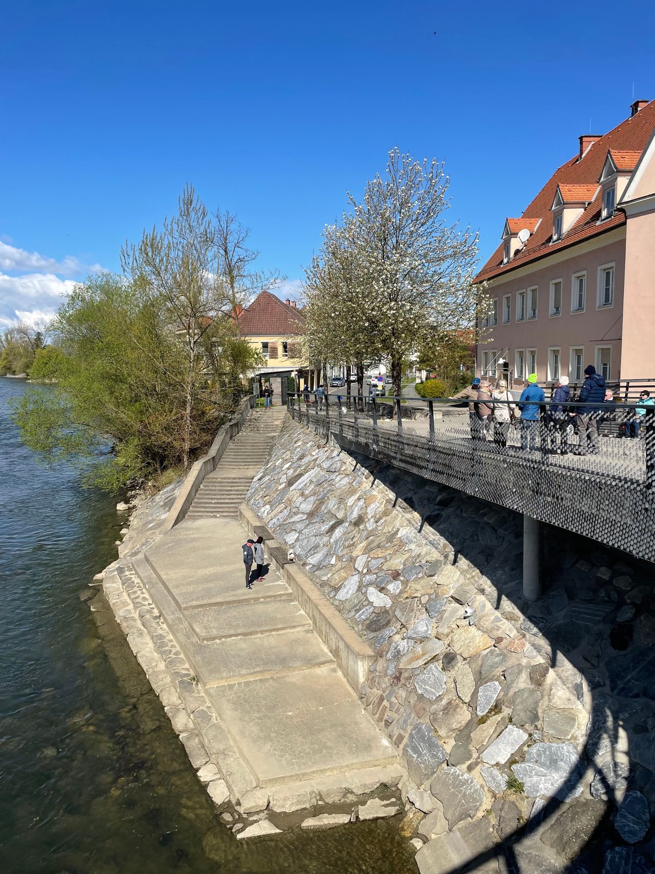 Eine Betontreppe führt hinunter zum Ufer eines Gewässers, an dem eine Person geht, während sich Menschen auf einer Metallbrücke versammeln, die einen Fluss mit Bäumen und Gebäuden im Hintergrund überblickt.