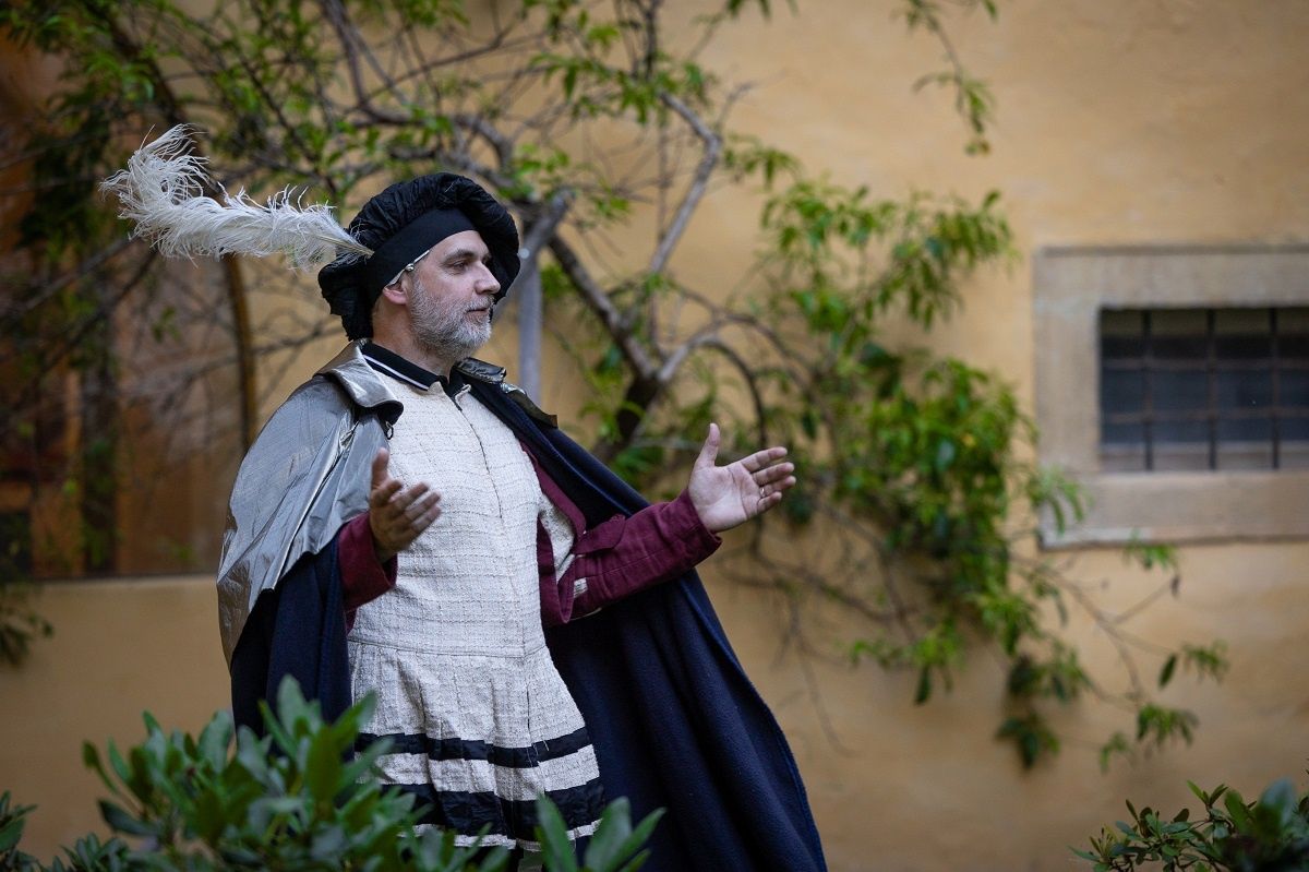 Ein Mann in einem Kostüm mit Federhut und Umhang deutet mit beiden Händen vor einer Wand.