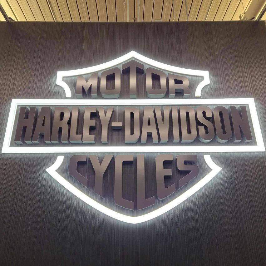Das Harley-Davidson Cycles-Logo wird an einer Wand mit Metallfinish gezeigt. Das Logo ist von weißen Lichtern beleuchtet, was einen auffälligen Kontrast erzeugt.