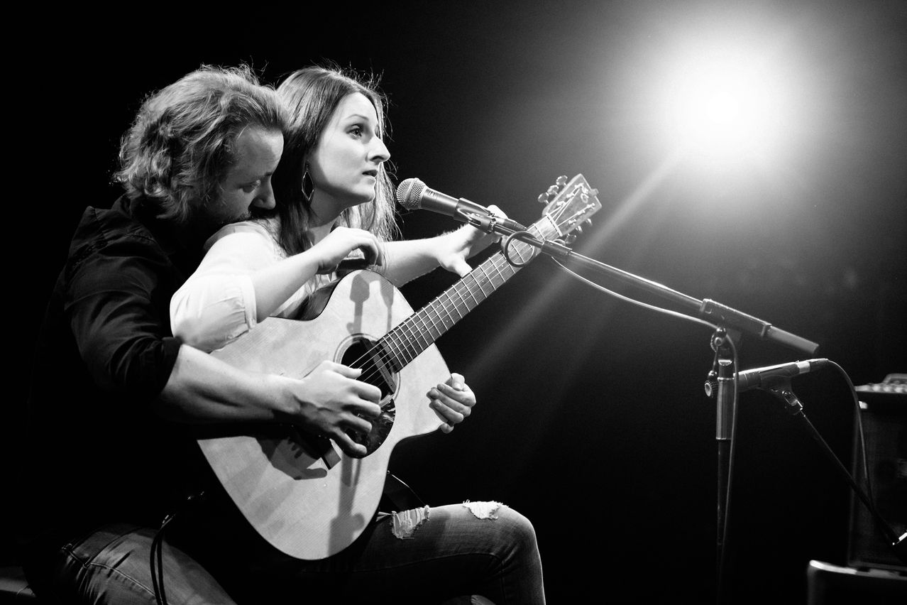 Ein Schwarz-Weiß-Bild einer Frau, die singt, während ein Mann neben ihr Gitarre spielt, beide schauen einander an, vor einem Mikrofon.