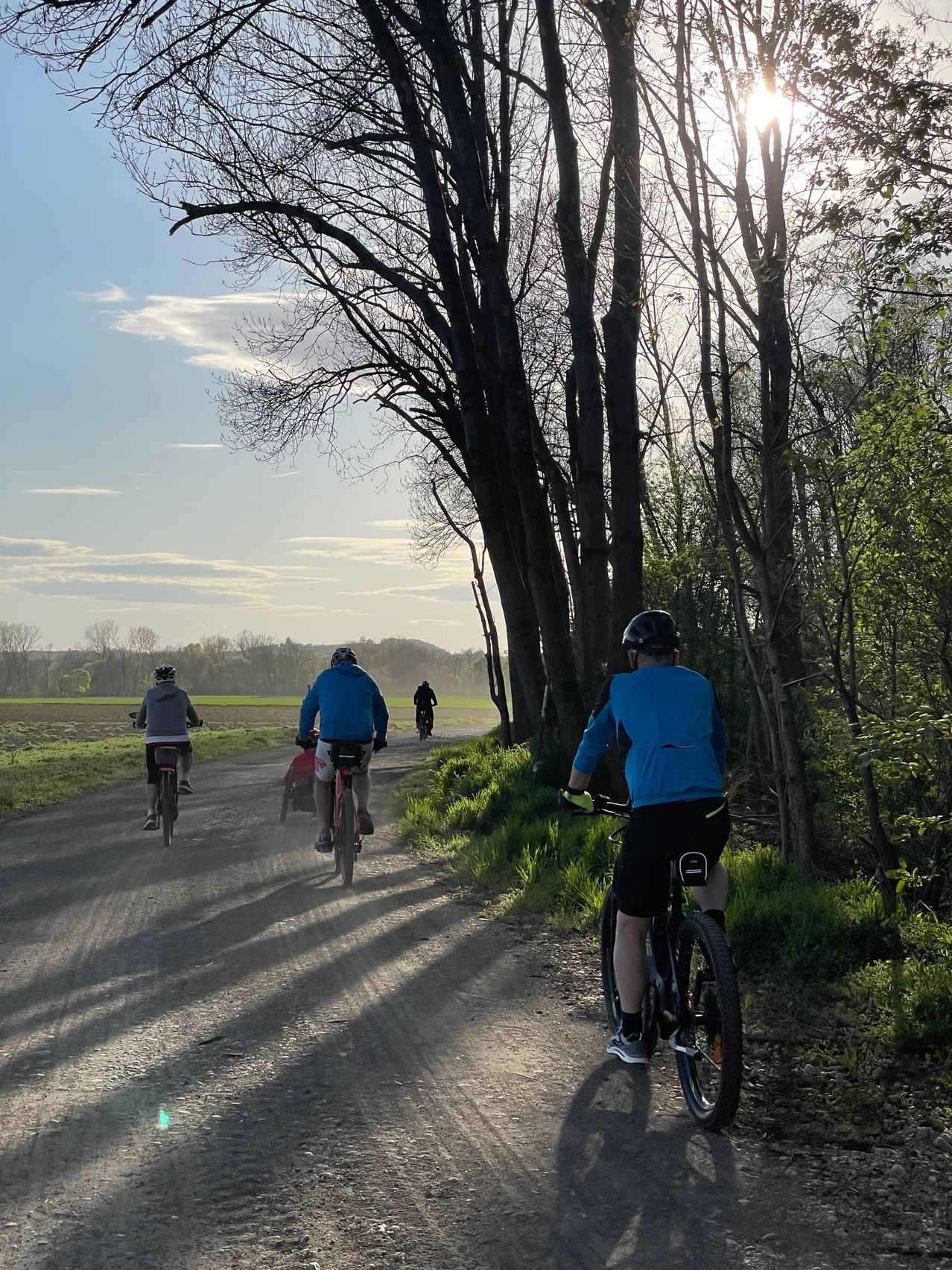 Eine Gruppe von Radfahrern fährt auf einem Feldweg, der von Bäumen gesäumt ist. Der Himmel ist klar mit einigen Wolken. Ein Radfahrer hat ein Kind auf seinem Fahrrad.