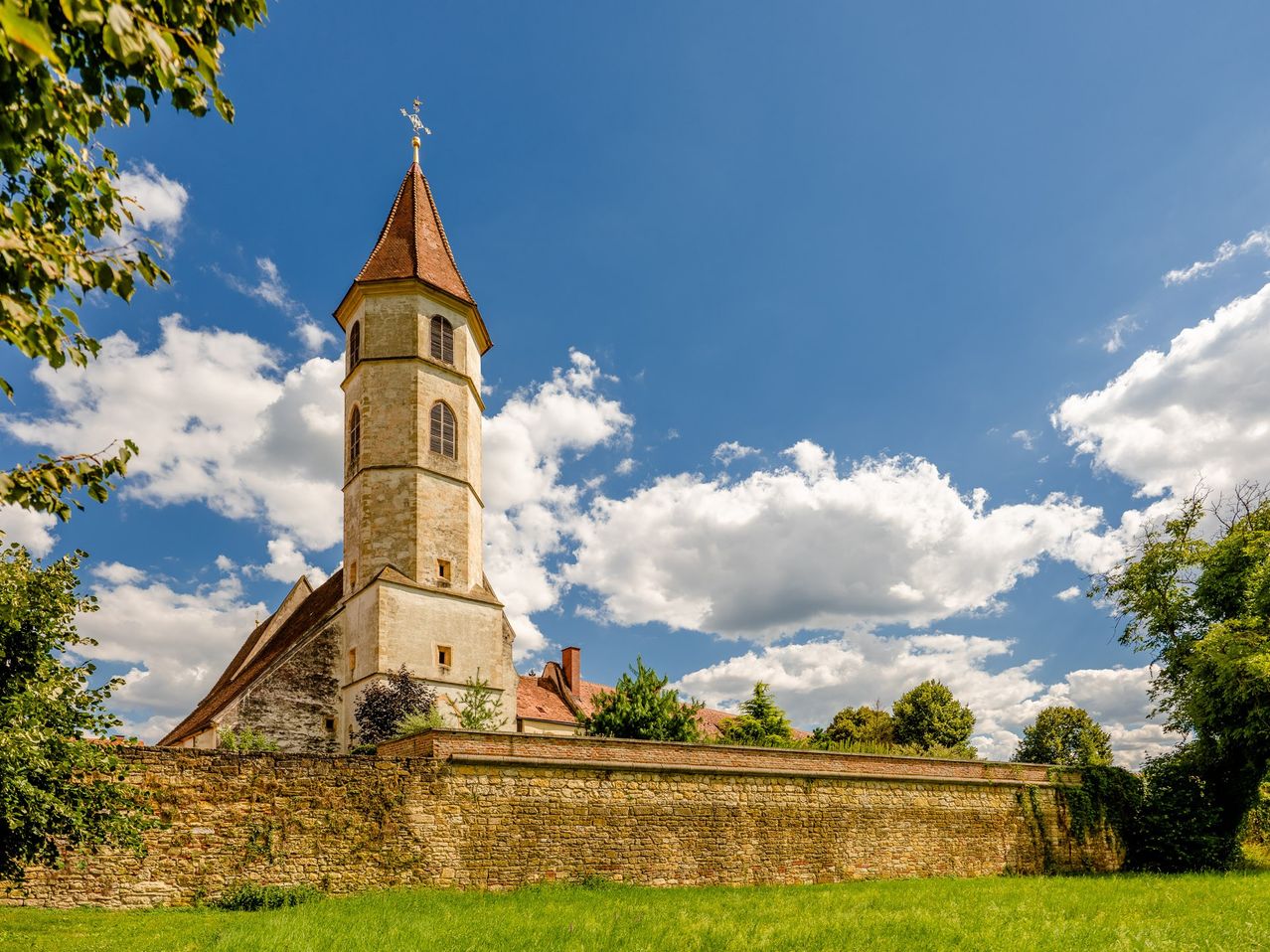 Eine alte Steinkirche mit einem hohen Turm steht auf einem Feld, umgeben von einer Steinmauer. Der Turm hat eine Wetterfahne auf der Spitze. Der Himmel ist blau mit verstreuten Wolken.