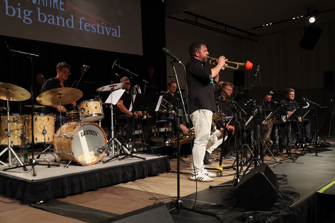 Eine Band tritt auf der Bühne auf, mit einem Trompeter im Stehen und anderen sitzend. Trommeln, Becken und verschiedene Instrumente sind sichtbar. Eine große Leinwand im Hintergrund zeigt 'RE and festival.'