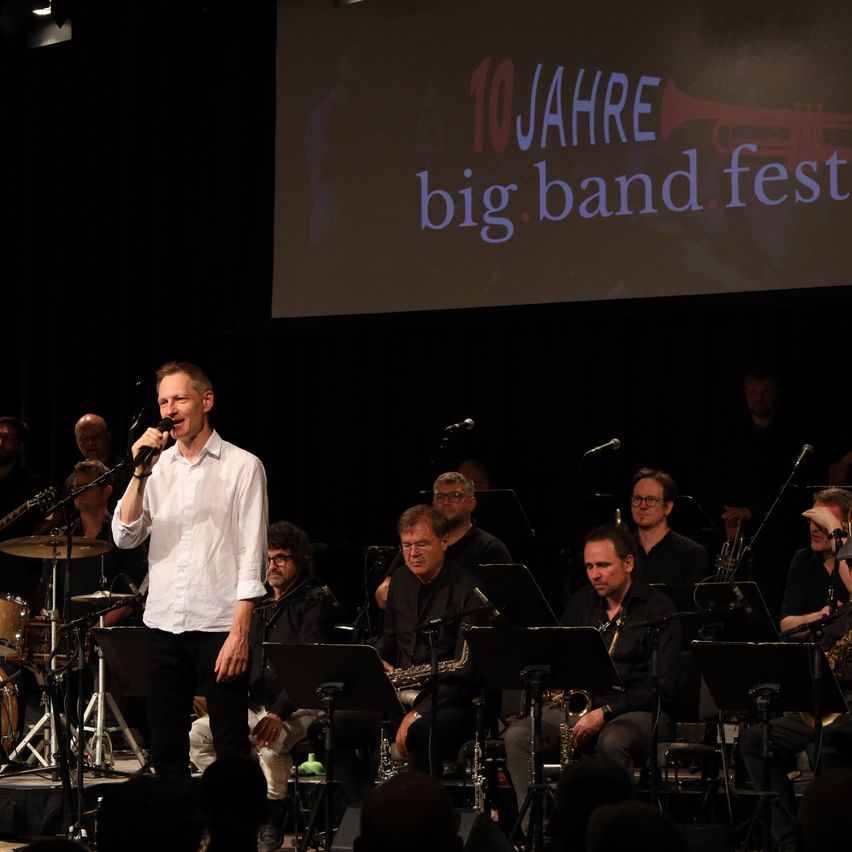 Eine Band tritt auf der Bühne auf, mit einer großen Leinwand im Hintergrund, auf der die Worte '10 Jahre big band fest' stehen. Der Leadsänger steht vorne mit einem Mikrofon. Dahinter spielen andere Bandmitglieder verschiedene Instrumente.