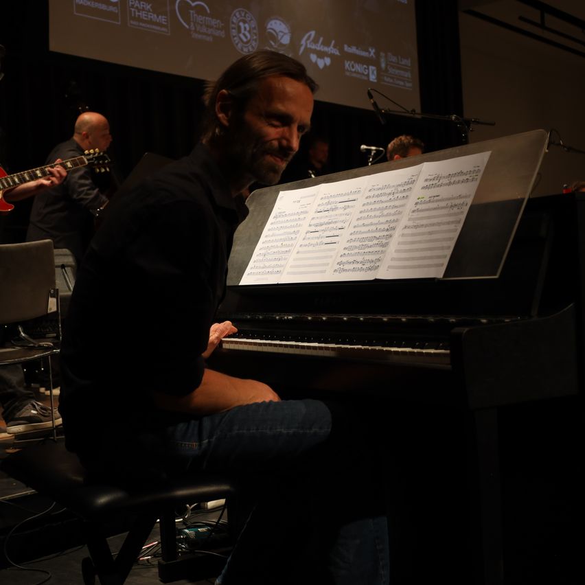 Ein Musiker spielt Klavier mit Notenblättern vor sich auf der Bühne. Hinter ihm spielt eine andere Person Gitarre. Im Hintergrund zeigt eine Projektionsleinwand Logos.