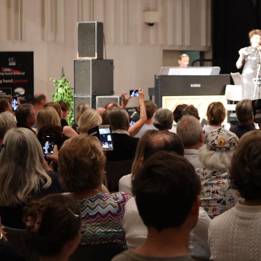 Eine Menschenmenge sitzt vor einer Bühne, auf der eine Frau in ein Mikrofon singt. Die Zuschauer halten ihre Telefone hoch und machen Fotos.
