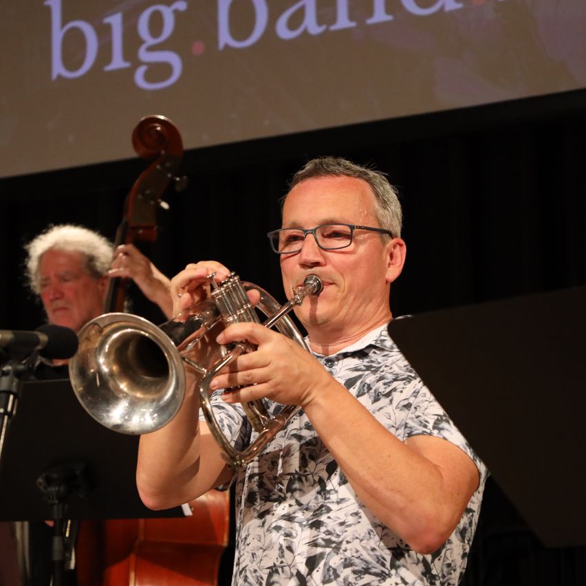 Ein Mann spielt eine Trompete während einer Aufführung, mit einer großen Leinwand im Hintergrund, auf der 'Big Band' steht. Ein anderer Musiker spielt Cello.