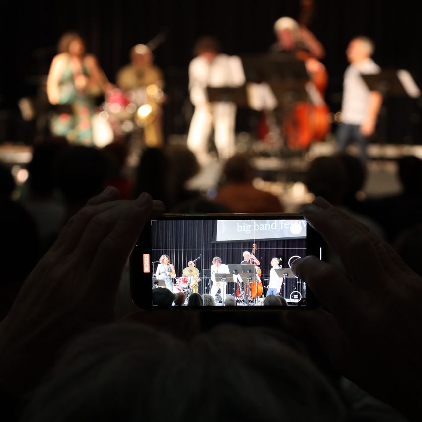 Eine Person macht ein Foto einer Big-Band-Aufführung mit ihrem Handy, das eine verschwommene Bühne mit Musikern zeigt, die Instrumente spielen.