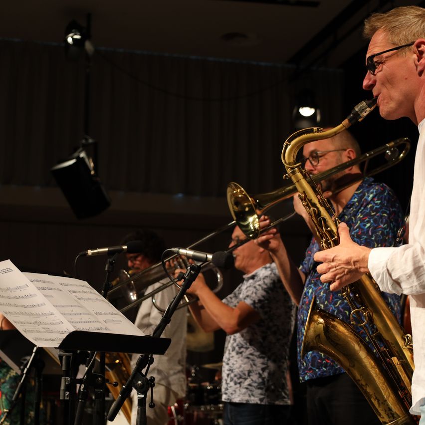 Eine Band probt mit einem Saxophonisten im Vordergrund, einem Posaunisten hinter ihm und einem Schlagzeuger im Hintergrund. Die Musiker sind von Bühnenlichtern beleuchtet.