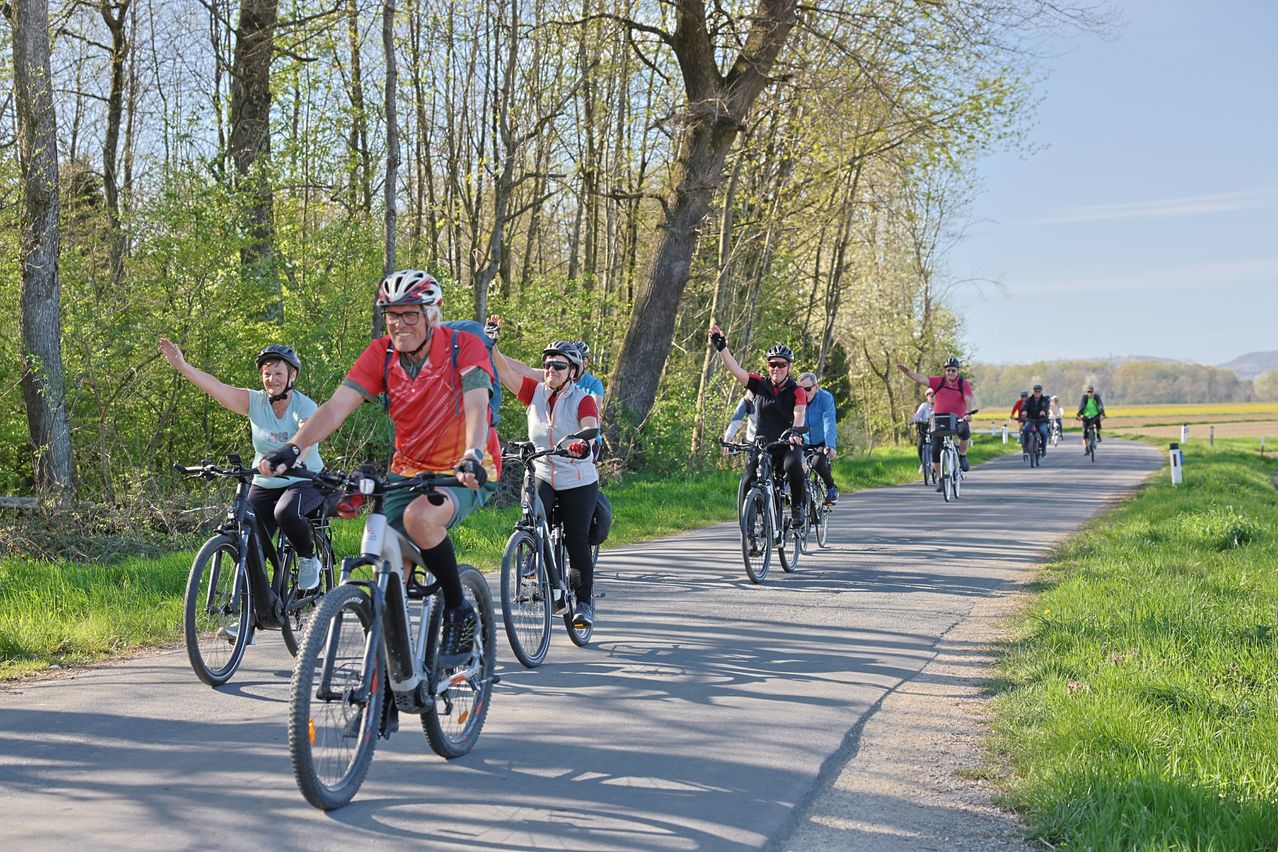 Eine Gruppe von Radfahrern fährt auf einer Straße in einem bewaldeten Gebiet, trägt Helme und Fahrradkleidung. Die Radfahrer sind in verschiedenen Positionen, einige stehen und heben ihre Hände. Die Straße hat grasige Seiten, und die Radfahrer scheinen die Fahrt zu genießen.