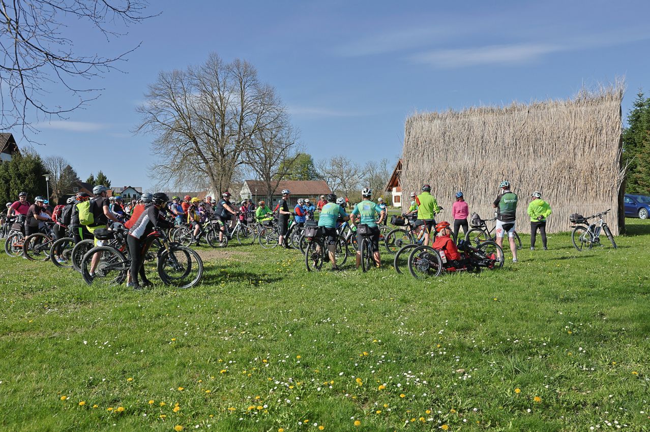 Eine Gruppe von Radfahrern versammelt sich auf einer Wiese. Sie tragen Helme und bunte Trikots. Eine Person in einem roten Hemd sitzt auf einem Dreirad. Dahinter sind ein strohgedecktes Gebäude und Bäume zu sehen.