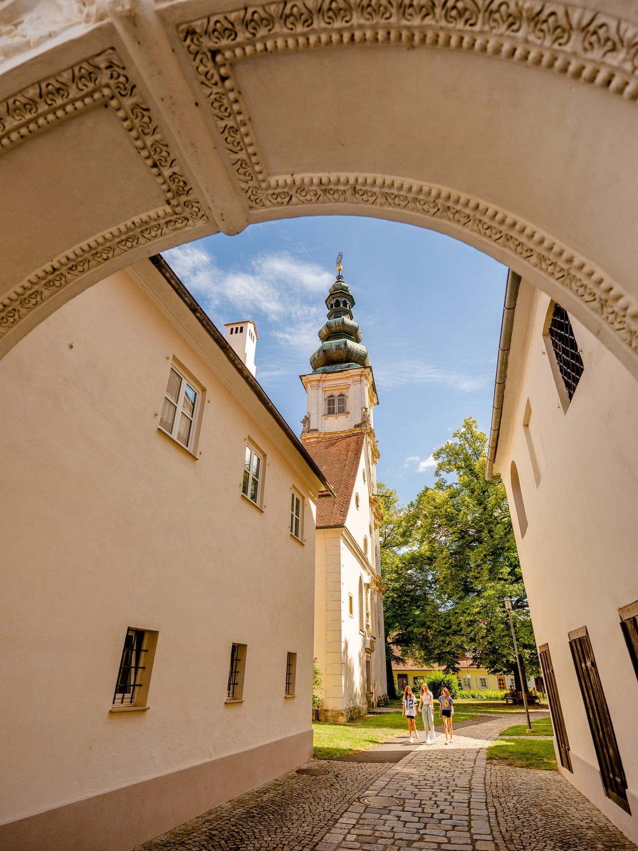 Ein Blick durch einen Bogen zeigt eine Kirche mit einem Turm, vor der drei Menschen an einem sonnigen Tag gehen.