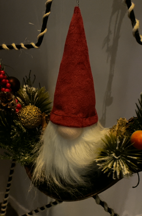 Ein roter und weißer Weihnachtszwerg, der an einer Schnur hängt, umgeben von grünen Blättern und roten Beeren.