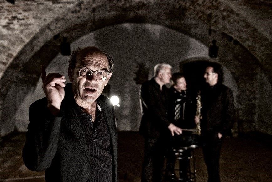 Ein älterer Mann in einem schwarzen Anzug und einer Brille macht eine Geste mit seiner Hand, während zwei Männer mit Saxophonen hinter ihm in einem schwach beleuchteten Raum stehen.