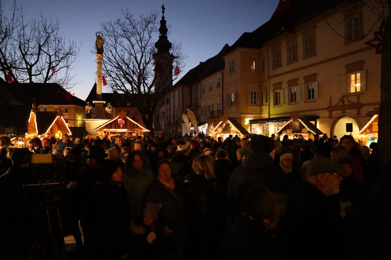 Eine Menschenmenge in Winterkleidung versammelt sich bei Dämmerung im Freien. Sie sind von beleuchteten Ständen umgeben.