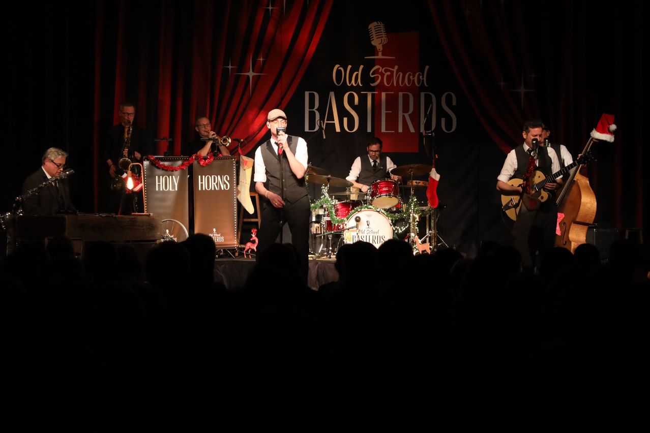 Eine Band tritt mit Mikrofon, Saxophon, Trompete und Schlagzeug auf der Bühne auf. Die Bühne hat einen Hintergrund mit dem Text Old School und BASIERDS.